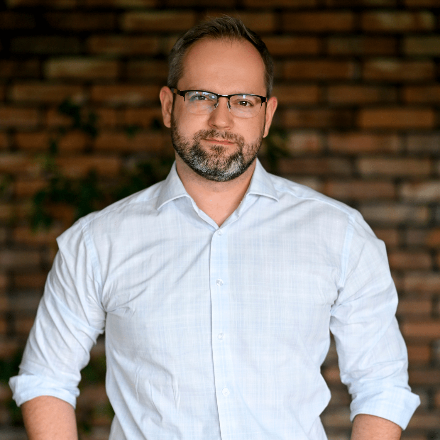 Mężczyzna w jasnej koszuli z rękawami podwiniętymi, stojący w pobliżu ceglanej ściany. Wygląda na pewnego siebie, z brodą i okularami, w otoczeniu zieleni. Idealny wizerunek dla producenta myjni bezdotykowych, reprezentujący profesjonalizm i zaufanie.