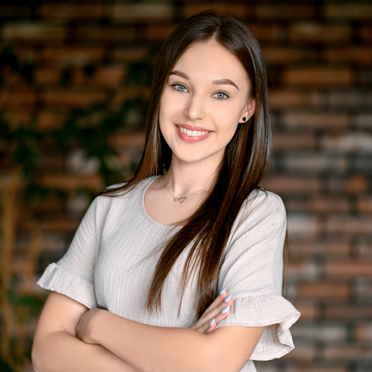 Młoda kobieta z długimi włosami i uśmiechem, ubrana w szaro-białą bluzkę, stoi z założonymi rękoma. W tle znajdują się ceglane ściany, co nadaje przytulny klimat. Idealna ilustracja dla producentów myjni bezdotykowych szukających pozytywnego wizerunku.