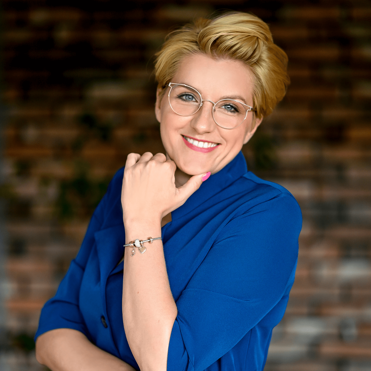 Młoda kobieta w niebieskiej bluzie z rękawami, z okularami i uśmiechem na twarzy, dotyka swojego podbródka. Tło w odcieniach brązu. Producent myjni bezdotykowych, profesjonalizm i pozytywna energia w obrazku.