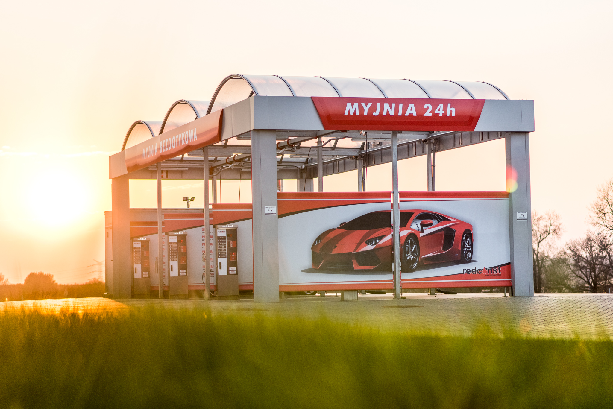 Nowoczesna myjnia bezdotykowa z widocznymi stanowiskami do mycia samochodów, z reklamą sportowego auta na ścianie. Obiekt czynny 24h, otoczony zielenią, przy zachodzie słońca, podkreślający komfort korzystania z myjni dla kierowców.