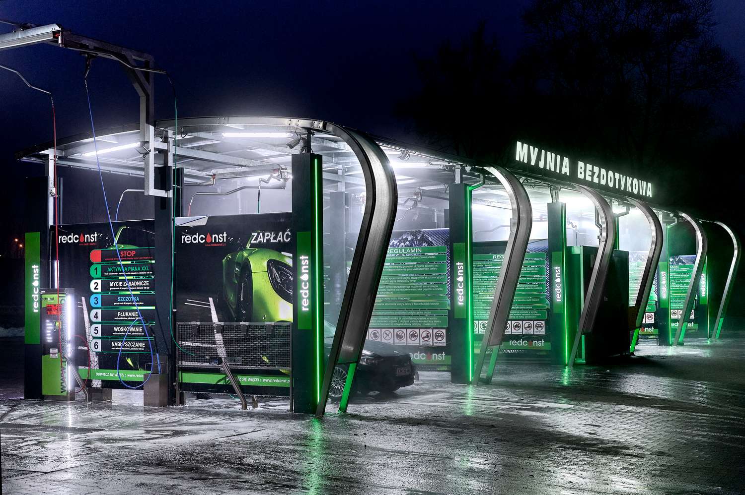 Nowoczesna myjnia bezdotykowa z jasnym oświetleniem, widoczna w nocy. Na ścianach umieszczone instrukcje dotyczące mycia samochodów oraz cen usług. Całość utrzymana w zielono-czarnych kolorach, z wyraźnym napisem "MYJNIA BEZDOTYKOWA". Idealne rozwiązanie dla posiadaczy aut.