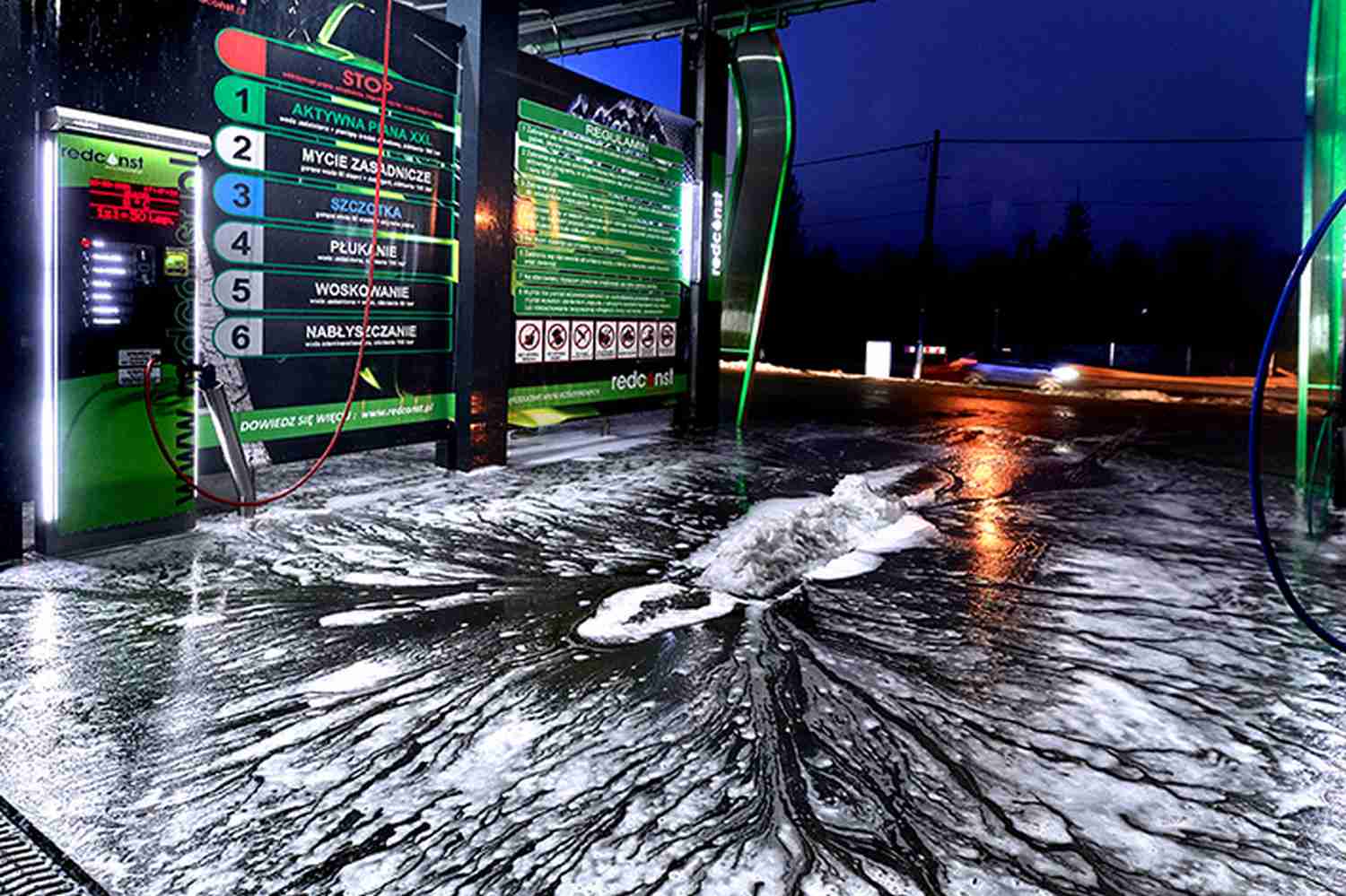 Bezdotykowa myjnia samochodowa w nocy, z widoczną pianą na podłodze oraz wyświetlaczem instrukcji mycia. W tle widać oświetlone elementy myjni oraz pojazdy na drodze, co podkreśla nowoczesne rozwiązania w branży myjni bezdotykowych.