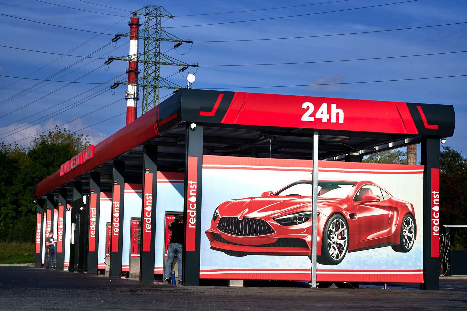 Nowoczesna myjnia bezdotykowa działająca 24 godziny na dobę, z dużymi reklamami pokazującymi eleganckie samochody. W tle widoczne instalacje elektryczne i zieleń, co tworzy nowoczesną i funkcjonalną przestrzeń do mycia pojazdów. Idealna oferta od producenta myjni bezdotykowej.