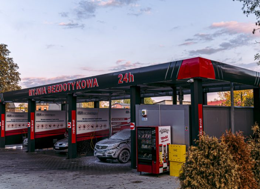 Myjnia bezdotykowa czynna 24h, z nowoczesnymi stanowiskami do mycia samochodów. W tle widać pojazdy pokryte pianą, a z przodu automaty z napojami. Idealne miejsce dla osób szukających wygodnej myjni bezdotykowej.