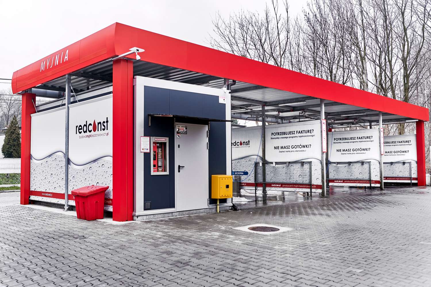 Nowoczesna myjnia bezdotykowa z czerwonym dachem, zawierająca automatyczny terminal i informacje o płatnościach. W tle widoczne banery reklamowe. Idealna lokalizacja dla kierowców szukających wysokiej jakości mycia samochodu. Redconst jako producent myjni bezdotykowych.