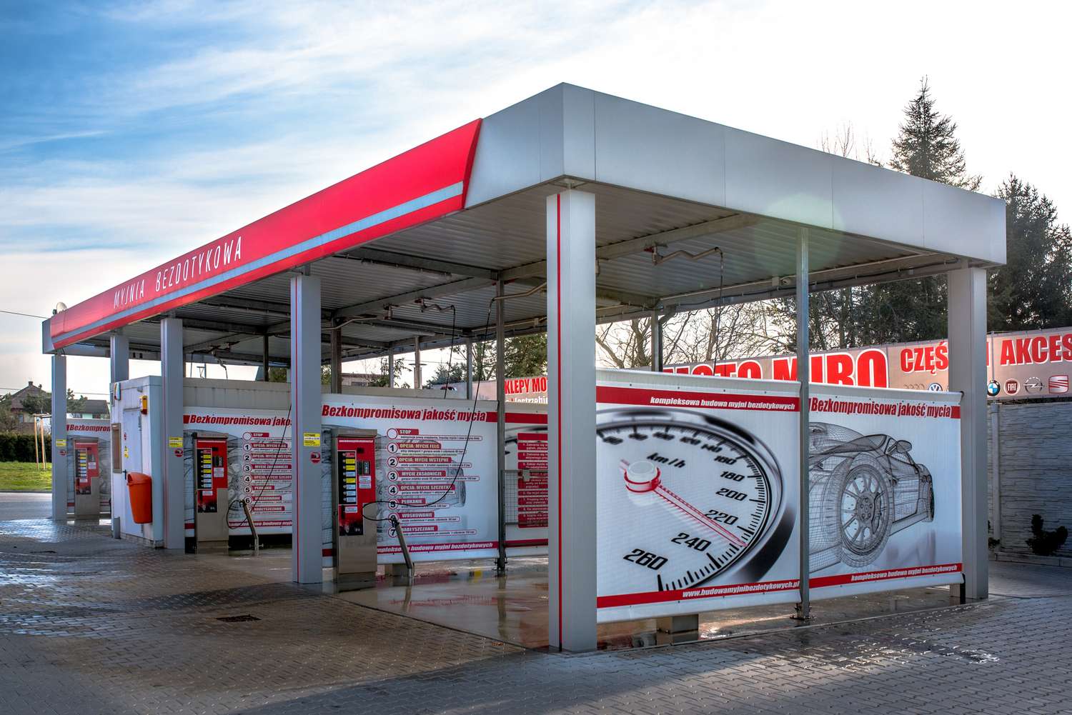 Myjnia bezdotykowa z nowoczesnym wyposażeniem, prezentująca różne usługi mycia samochodów. W tle widoczne grafiki i instrukcje dotyczące obsługi, z jasnym niebieskim niebem. Idealna dla producentów myjni bezdotykowych poszukujących inspiracji.