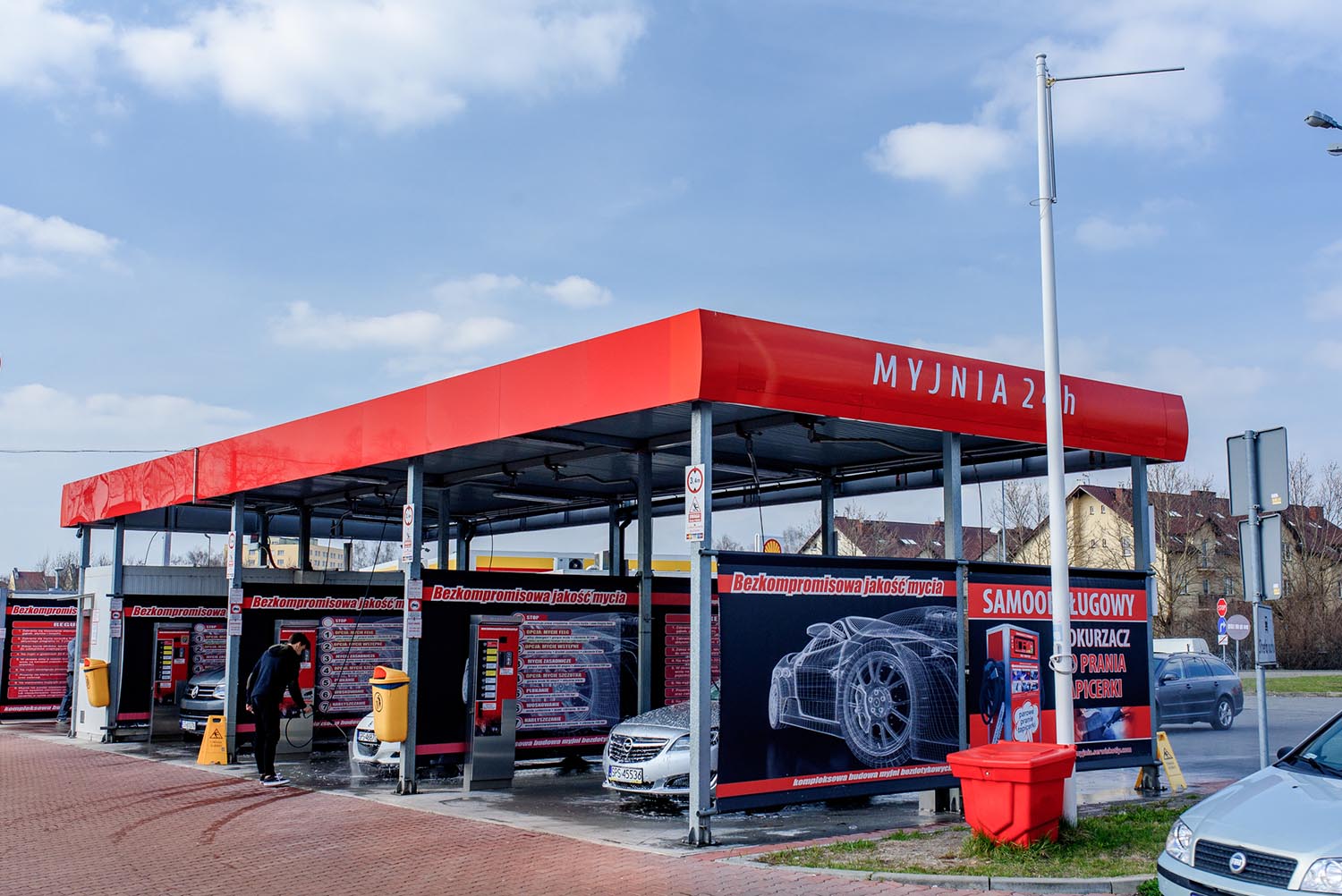 Myjnia bezdotykowa z wieloma stanowiskami do mycia samochodów, z nowoczesnym wyposażeniem i reklamami podkreślającymi najwyższą jakość usług. W tle widać błękitne niebo i okoliczny krajobraz.