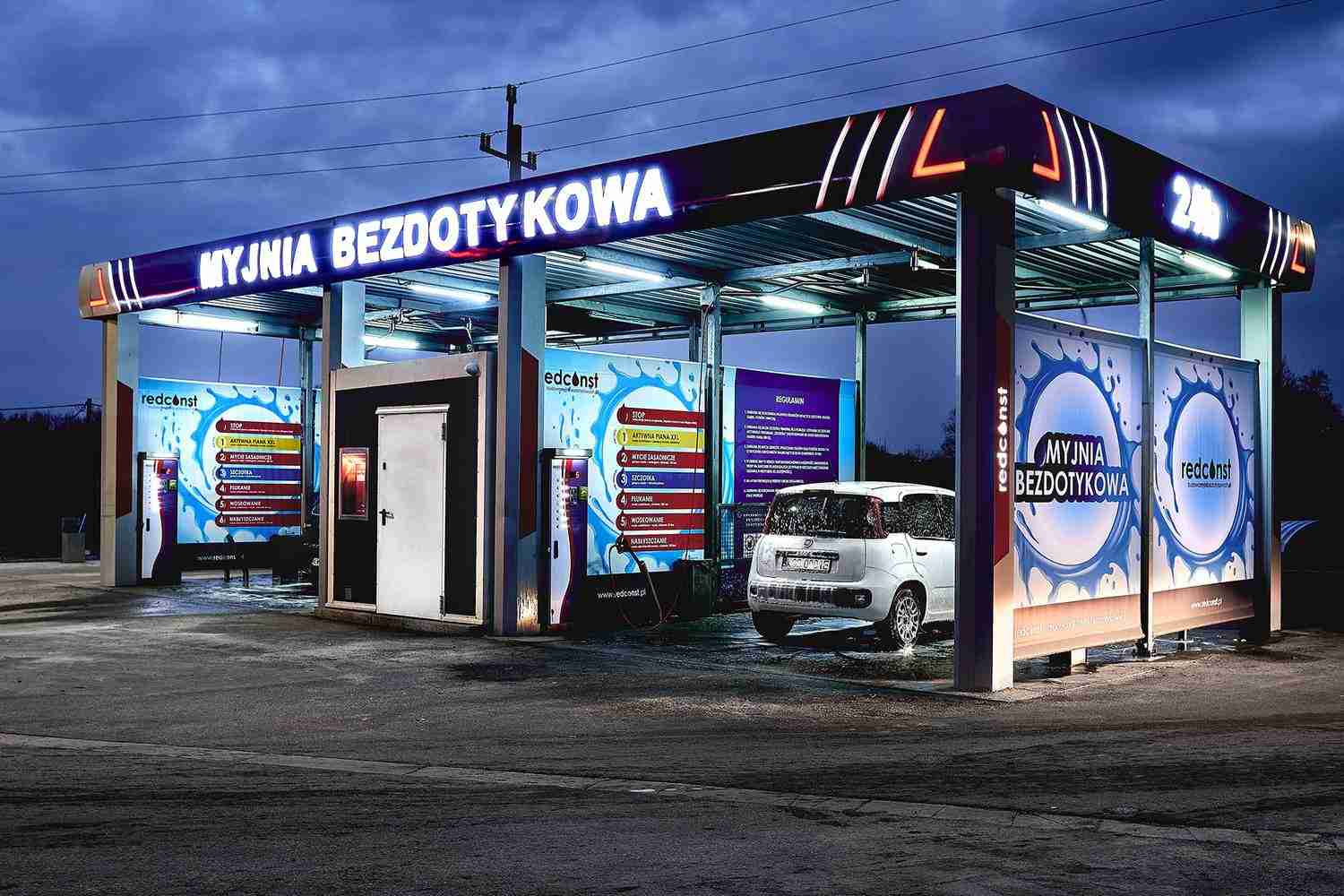 Bezdotykowa myjnia samochodowa z logo "MYJNIA BEZDOTYKOWA", podświetlona w nocy. W tle widać instrukcje użytkowania oraz przygotowane do mycia auto. Idealne miejsce dla kierowców poszukujących efektywnego czyszczenia pojazdów.