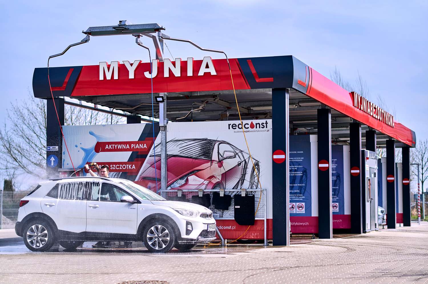 Myjnia bezdotykowa z aktywną pianą, gdzie mężczyzna myje biały samochód. W tle widoczne elementy reklamowe i instrukcje dotyczące korzystania z myjni. To nowoczesne rozwiązanie dla właścicieli pojazdów, poszukujących skutecznego i wygodnego mycia.