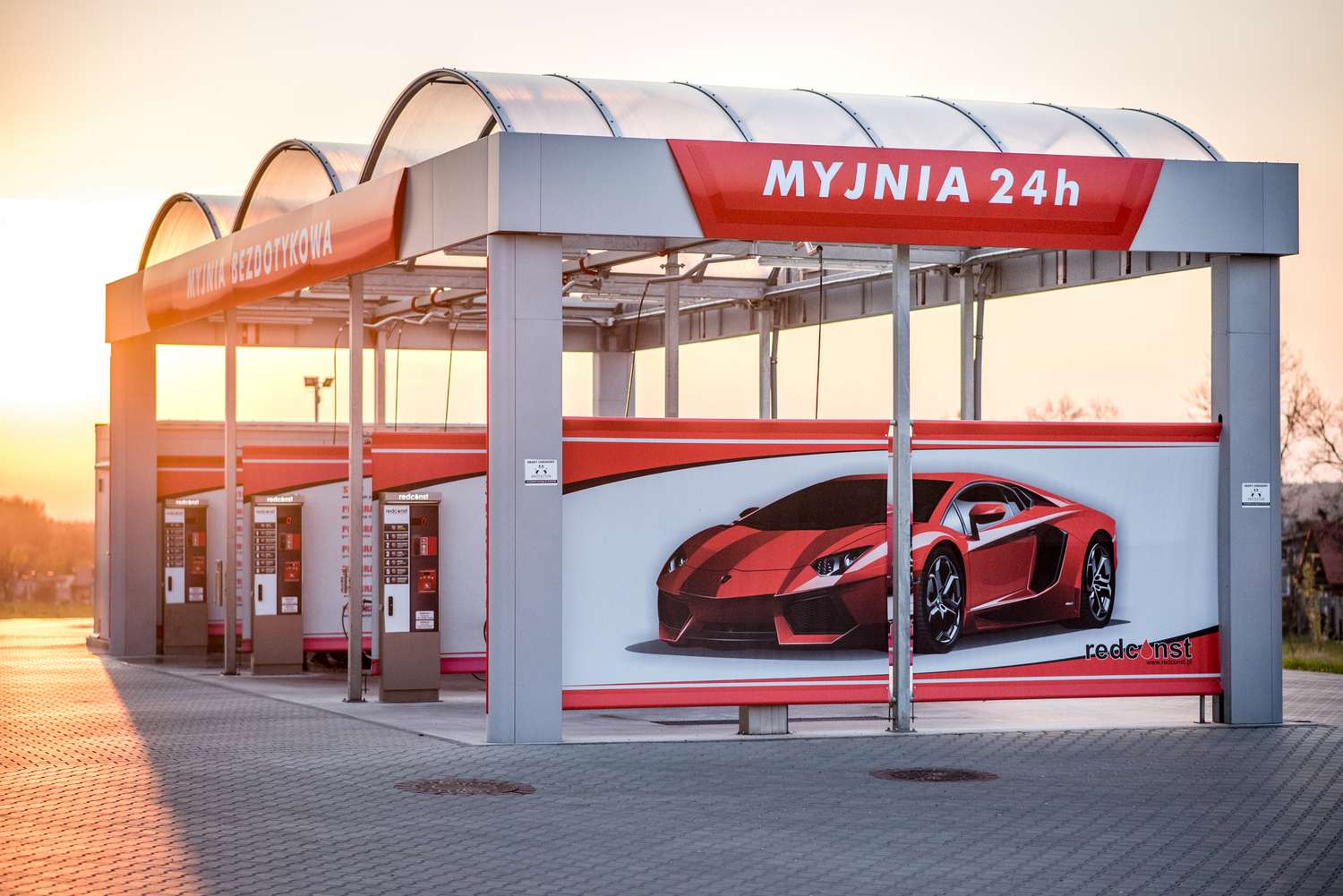 Nowoczesna myjnia bezdotykowa 24h z eleganckim stoiskiem i zimnym oświetleniem. Na ścianie zewnętrznej znajduje się grafika sportowego samochodu, co podkreśla luksusowy charakter usługi. Myjnia idealna dla właścicieli cennych aut, oferująca wygodę i efektywność czyszczenia.