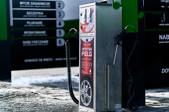 Stacja myjni bezdotykowej z metalowym urządzeniem do czyszczenia felg. Na panelu widoczne przyciski oraz instrukcje użytkowania. W tle tablica z numerami usług mycia. Idealne dla producentów myjni bezdotykowych.