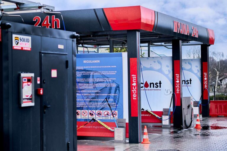 Zewnętrzny widok myjni bezdotykowej działającej 24 godziny na dobę, z regulaminem oraz logo producenta myjni bezdotykowych. Przed myjnią znajdują się czerwone pachołki i czarna szafa zabezpieczeń.