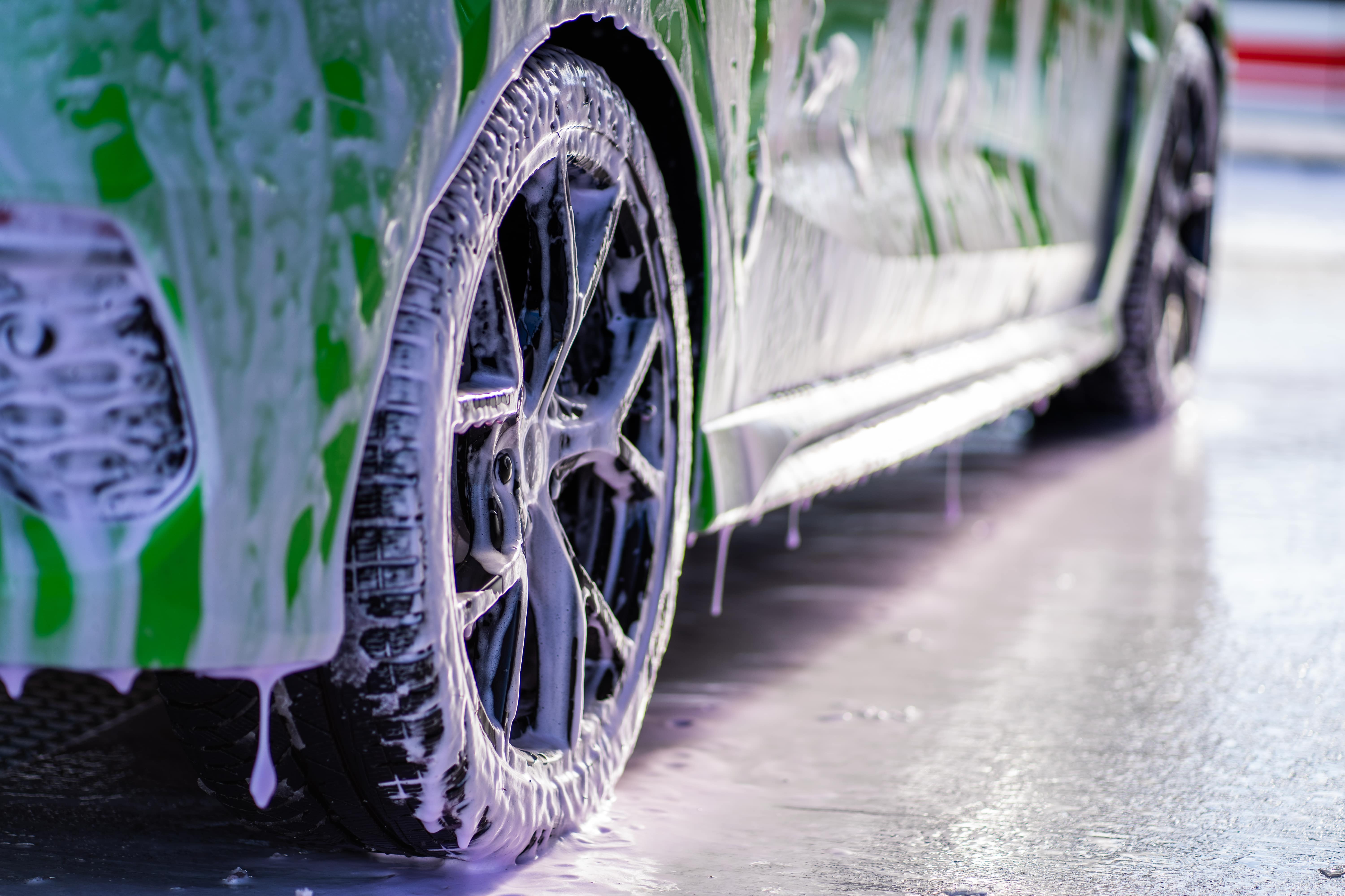 Zbliżenie na koło samochodu pokryte bąbelkami aktywnej piany w myjni bezdotykowej. Zielony lakier pojazdu lśni, a z boku spływa piana, tworząc efektowny widok. Idealne dla producentów myjni bezdotykowych, prezentując skuteczność mycia i pielęgnacji samochodów.